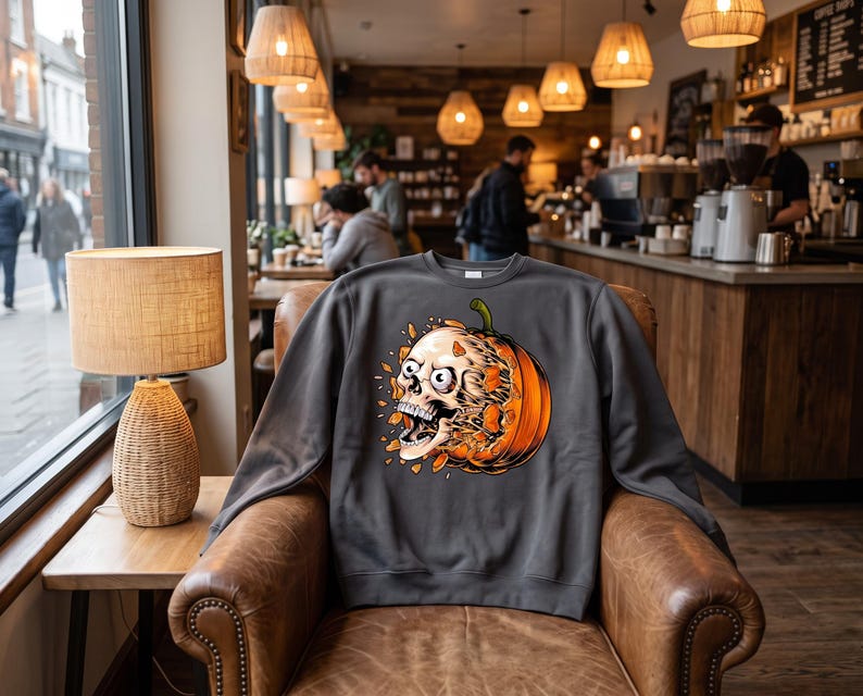 May include: A charcoal grey crewneck sweatshirt featuring a graphic print of a white human skull emerging from a bright orange pumpkin. The skull has wide, startled eyes and an open mouth, with orange pumpkin fragments floating around the design.