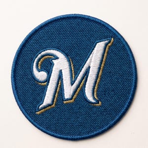 May include: A circular embroidered patch featuring a dark blue background with a stylized white letter M in the center. The letter M has a thin gold outline and a decorative swirled tail on the left side. The patch has a dark blue stitched border.