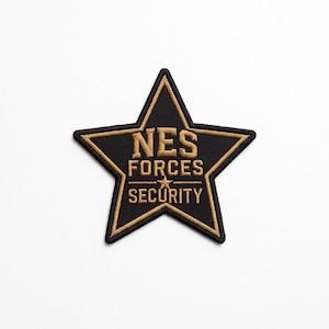 Könnte beinhalten: Ein sternförmiger Aufnäher mit schwarzem Hintergrund und goldfarbenem Rand. Der Text NES FORCES SECURITY ist in goldfarbenem Garn in die Mitte gestickt, wobei ein kleiner goldener Stern die Wörter FORCES und SECURITY trennt.