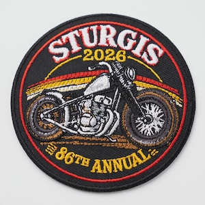 Puede incluir: Un parche bordado circular con fondo negro que presenta un gráfico de una motocicleta blanca. El texto STURGIS 2026 aparece en la parte superior y 86TH ANNUAL en la inferior con letras amarillas y blancas. Líneas rojas y amarillas decoran el diseño.