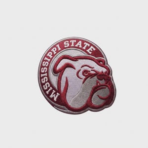 Puede incluir: Un parche bordado circular que presenta un gráfico de cabeza de bulldog en color granate y blanco. El texto MISSISSIPPI STATE está bordado en letras mayúsculas blancas a lo largo del borde superior e izquierdo del marco circular granate.