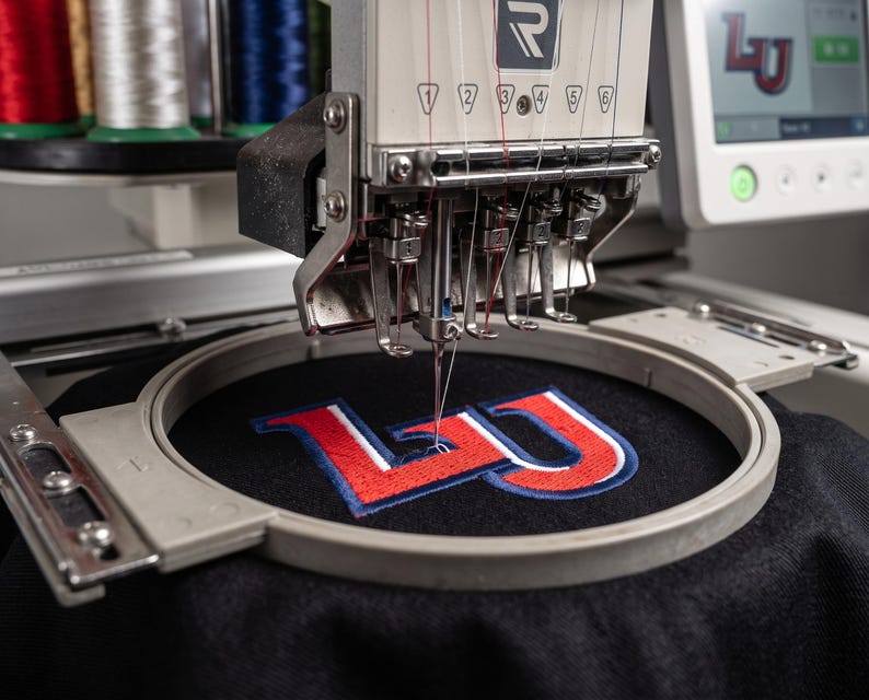 May include: A multi-needle embroidery machine stitching a red, white, and blue LU logo onto black fabric held in a circular hoop. The machine features a digital screen displaying the logo and multiple spools of thread in the background.