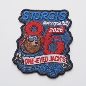 May include: An embroidered patch featuring a brown bear wearing sunglasses and a stars and stripes bandana. The text reads STURGIS Motorcycle Rally 2026 86th Anniversary ONE-EYED JACK'S Saloon. The design uses red, blue, white, and brown thread.