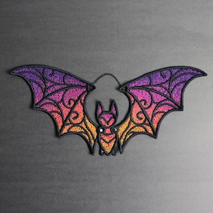 May include: An embroidered bat patch featuring a gradient color scheme that transitions from purple at the top of the wings to orange at the bottom. The design includes intricate black stitched filigree patterns and a thin black chain loop.