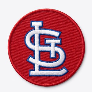 May include: A circular embroidered patch featuring a red background with a white and blue interlocking STL logo in the centre. The patch has a thick red stitched border and a textured fabric surface, showcasing detailed thread work throughout the design.