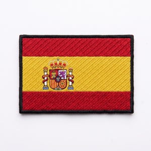 May include: A rectangular embroidered patch featuring the flag of Spain with horizontal red and yellow stripes and the national coat of arms centered on the left side, all enclosed within a thick black stitched border on a white background.