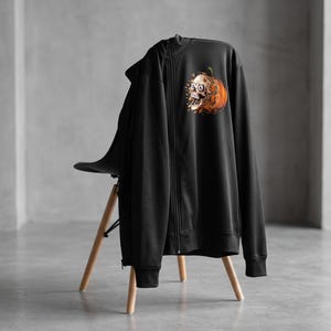 May include: A black zip-up hoodie featuring a graphic print on the upper left chest. The design depicts a human skull emerging from an orange pumpkin, with small orange leaf-like fragments scattered around the base of the pumpkin illustration.