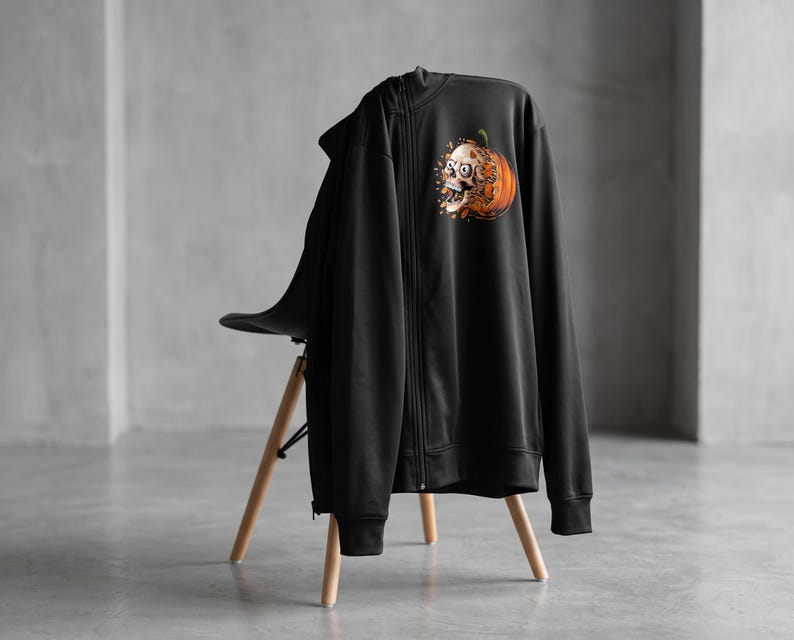 May include: A black zip-up hoodie featuring a graphic print on the upper left chest. The design depicts a human skull emerging from an orange pumpkin, with small orange leaf-like fragments scattered around the base of the pumpkin illustration.