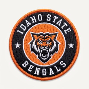 May include: A circular embroidered patch featuring an orange tiger head at the center on an orange background. A black border surrounds the center, containing the white text IDAHO STATE BENGALS and two white stars on the left and right sides.