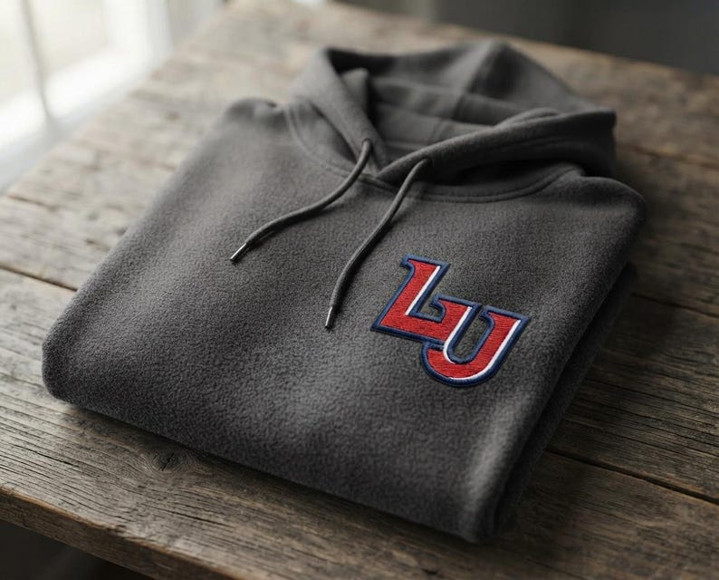 May include: A charcoal gray fleece hoodie featuring a hood with matching drawstrings and a prominent embroidered patch on the chest. The patch displays the letters LU in bold red with white and navy blue outlines, stitched onto the fabric.