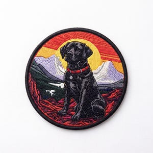 May include: A circular embroidered patch featuring a black Labrador retriever wearing a red collar. The dog sits in front of a landscape with purple mountains, a yellow sun, and a red sky, all enclosed within a thick black stitched border.