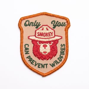Puede incluir: Un parche bordado en forma de escudo con fondo beige y borde naranja. Presenta un oso rojo con un sombrero beige que lleva la palabra SMOKEY. El texto dice Only You CAN PREVENT WILDFIRES en letras bordadas de color verde oscuro.