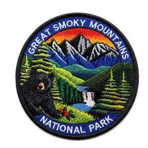Pode incluir: Um emblema bordado circular apresentando um urso preto em primeiro plano, uma cachoeira, pinheiros verdes e montanhas azuis sob um céu ao pôr do sol. O texto GREAT SMOKY MOUNTAINS NATIONAL PARK está bordado em branco na borda azul.