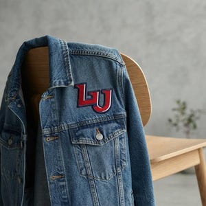May include: A blue denim jacket featuring a prominent embroidered patch on the chest. The patch displays the letters LU in bold red with a white and dark blue outline. The jacket includes a classic collar, metal buttons, and two chest pockets.