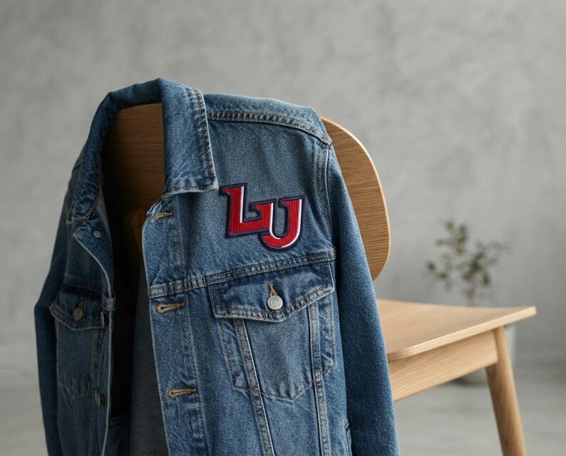 May include: A blue denim jacket featuring a prominent embroidered patch on the chest. The patch displays the letters LU in bold red with a white and dark blue outline. The jacket includes a classic collar, metal buttons, and two chest pockets.