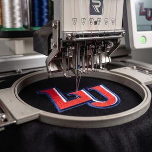 May include: A multi-needle embroidery machine stitching a red, white, and blue LU logo onto black fabric held in a circular hoop. The machine features a digital screen displaying the logo and multiple spools of thread in the background.