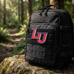 May include: A black tactical backpack featuring a red and white embroidered patch with the letters LU on the front. The bag includes multiple zippered compartments, adjustable side compression straps with plastic buckles, and a modular webbing system.