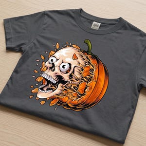May include: A dark grey short-sleeve t-shirt featuring a graphic print of a human skull bursting out from the centre of a bright orange pumpkin. The skull has wide eyes and an open mouth, with orange pumpkin shards flying outward in all directions.