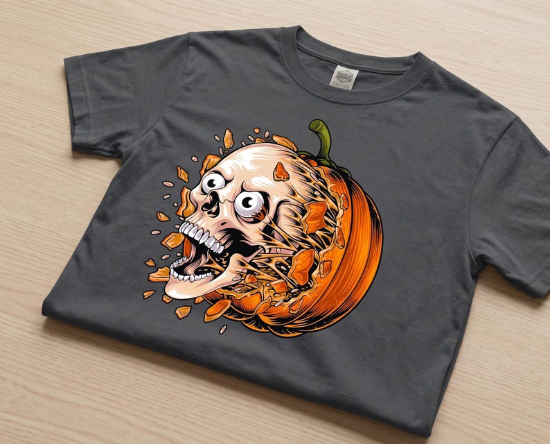 May include: A dark grey short-sleeve t-shirt featuring a graphic print of a human skull bursting out from the centre of a bright orange pumpkin. The skull has wide eyes and an open mouth, with orange pumpkin shards flying outward in all directions.