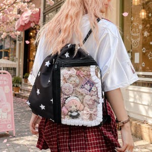 Könnte beinhalten: Ein schwarzer Kordelzug-Rucksack mit weißem Sternenmuster und einer transparenten Fronttasche. Die Tasche ist mit dekorativen Gegenständen gefüllt, darunter eine kleine Plüschpuppe, mehrere runde Anstecknadeln, rechteckige Karten und rosa Stoffschleifen.
