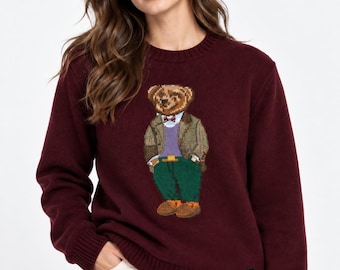May include: A long-sleeved, crew-neck sweater in a deep burgundy color. The front features a knitted graphic of a teddy bear wearing a green jacket, a purple sweater, a bow tie, and green trousers. A small black tag with the letters RL is on the hem.