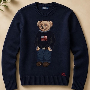 May include: A navy blue crewneck sweater featuring a knit graphic of a teddy bear wearing a dark sweater with an American flag design, blue jeans, and brown shoes. The Polo by Ralph Lauren label is at the neck, and RL is in red at the hem.