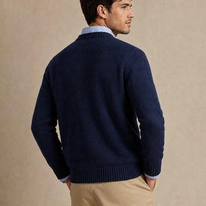 May include: A navy blue long-sleeved crewneck sweater with a ribbed collar, cuffs, and hem. The sweater is worn over a light blue collared button-down shirt, paired with tan trousers. The view shows the back of the garment.
