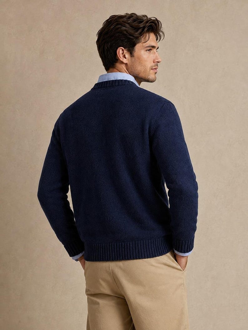 May include: A navy blue long-sleeved crewneck sweater with a ribbed collar, cuffs, and hem. The sweater is worn over a light blue collared button-down shirt, paired with tan trousers. The view shows the back of the garment.