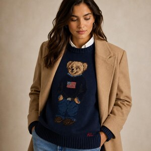 May include: A navy blue crewneck sweater featuring a knit graphic of a teddy bear wearing a sweater with an American flag design. The sweater has a ribbed hem with red RL embroidery and is layered over a white collared shirt under a tan coat.
