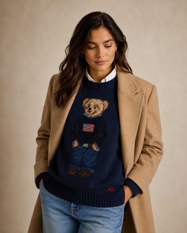 May include: A navy blue crewneck sweater featuring a knit graphic of a teddy bear wearing a sweater with an American flag design. The sweater has a ribbed hem with red RL embroidery and is layered over a white collared shirt under a tan coat.
