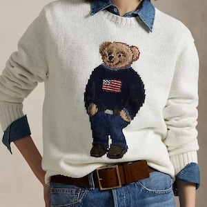 May include: A cream-colored long-sleeve knit sweater featuring a central graphic of a teddy bear wearing a navy blue sweater with an American flag design and blue jeans. The sweater is layered over a blue denim collared shirt and blue jeans.