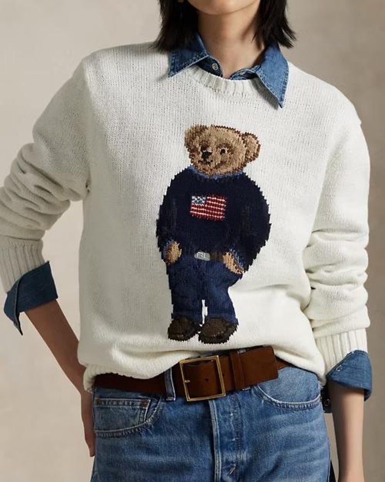May include: A cream-colored long-sleeve knit sweater featuring a central graphic of a teddy bear wearing a navy blue sweater with an American flag design and blue jeans. The sweater is layered over a blue denim collared shirt and blue jeans.