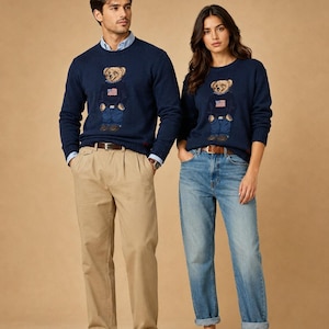 May include: Two navy blue crewneck sweaters featuring a central graphic of a teddy bear wearing a sweater with an American flag design. The sweaters are paired with light blue button-down shirts, khaki trousers, blue jeans, brown loafers, and white sneakers.