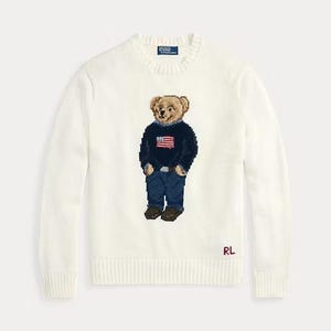 May include: A cream-colored long-sleeve crewneck sweater featuring a central intarsia-knit bear graphic. The bear wears a navy blue sweater with an American flag, blue jeans, and brown shoes. The Polo by Ralph Lauren label is at the neck.