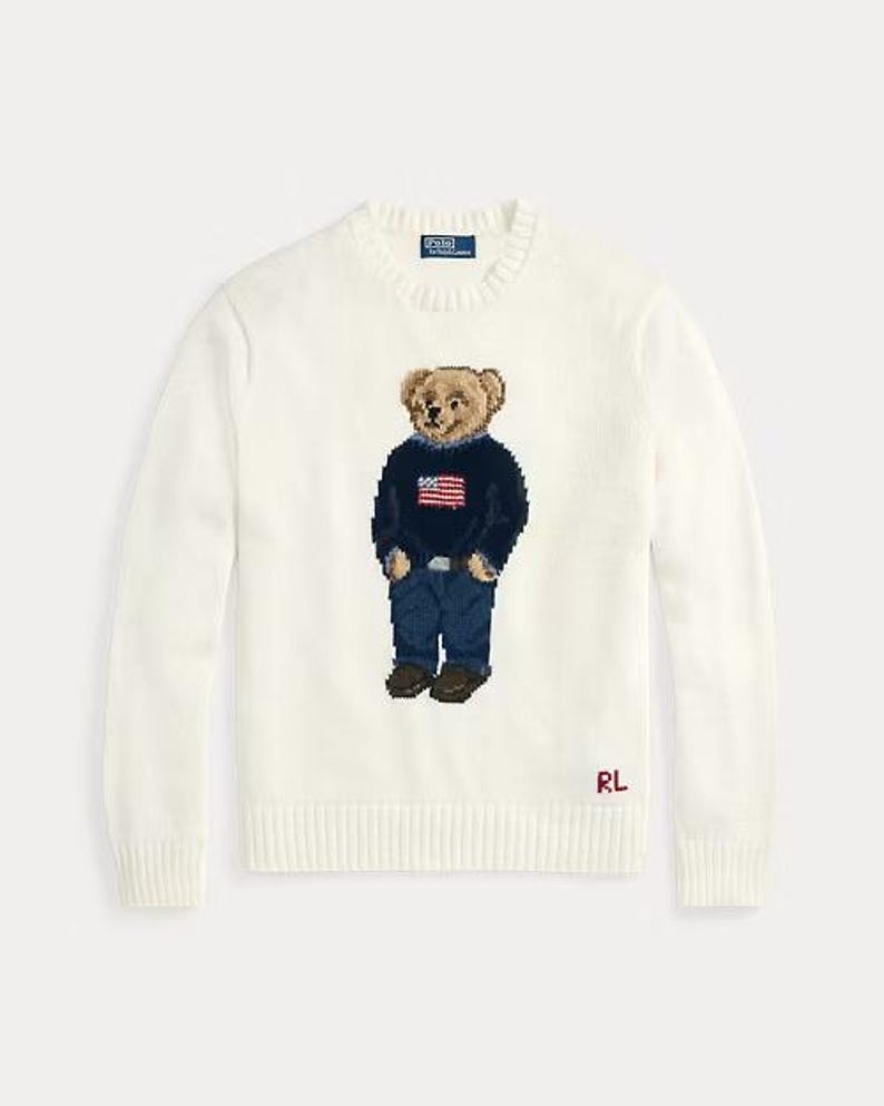 May include: A cream-colored long-sleeve crewneck sweater featuring a central intarsia-knit bear graphic. The bear wears a navy blue sweater with an American flag, blue jeans, and brown shoes. The Polo by Ralph Lauren label is at the neck.