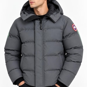 May include: A charcoal gray puffer jacket featuring a hood, a front zipper closure with a storm flap, and black ribbed cuffs. The jacket has a circular red, white, and blue logo patch on the upper left sleeve and horizontal quilted stitching throughout.