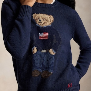 May include: A navy blue long-sleeve crewneck sweater featuring a knitted graphic of a teddy bear wearing a blue sweater with an American flag, blue jeans, and brown shoes. The letters RL are embroidered in red at the bottom right hem.