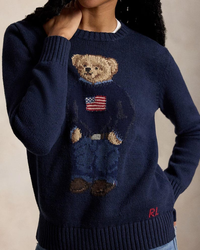 May include: A navy blue long-sleeve crewneck sweater featuring a knitted graphic of a teddy bear wearing a blue sweater with an American flag, blue jeans, and brown shoes. The letters RL are embroidered in red at the bottom right hem.
