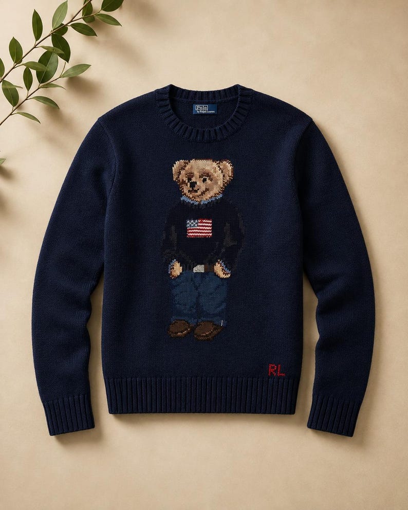 May include: A navy blue crewneck sweater featuring a knit graphic of a teddy bear wearing a dark sweater with an American flag design, blue jeans, and brown shoes. The Polo by Ralph Lauren label is at the neck, and RL is in red at the hem.