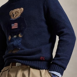May include: A navy blue long-sleeve crewneck sweater featuring a knitted bear graphic wearing a sweater with an American flag design. The sweater has a ribbed collar, cuffs, and hem, with the red embroidered letters RL near the bottom hem.
