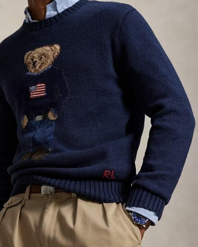 May include: A navy blue long-sleeve crewneck sweater featuring a knitted bear graphic wearing a sweater with an American flag design. The sweater has a ribbed collar, cuffs, and hem, with the red embroidered letters RL near the bottom hem.