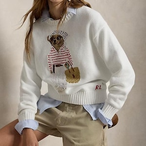 May include: A white long-sleeve knit sweater featuring a graphic of a bear wearing a polka dot headscarf, a red and white striped shirt, and white pants while carrying a tan woven bag. The letters RL are embroidered in red on the lower left.