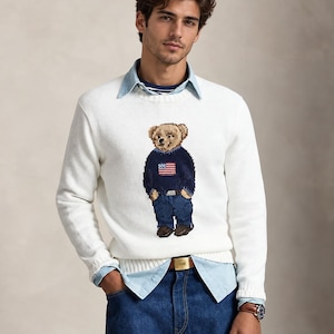May include: A white long-sleeved crewneck sweater featuring a central intarsia-knit bear graphic wearing a navy blue sweater with an American flag design, blue jeans, and brown shoes, layered over a light blue button-down shirt and blue jeans.