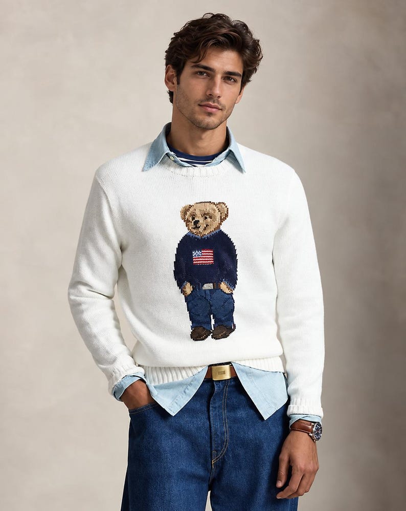 May include: A white long-sleeved crewneck sweater featuring a central intarsia-knit bear graphic wearing a navy blue sweater with an American flag design, blue jeans, and brown shoes, layered over a light blue button-down shirt and blue jeans.