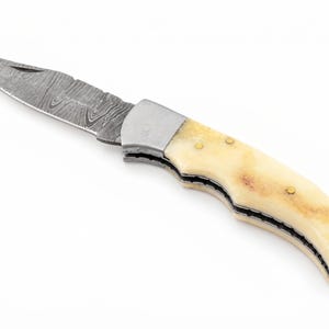Könnte beinhalten: Ein Klappmesser mit einer scharfen Klinge, die ein markantes, wellenförmiges Damaststahlmuster aufweist. Der Griff besteht aus glattem, cremefarbenem Knochenmaterial, das mit kleinen Messingstiften befestigt ist und eine Metallbacke besitzt.