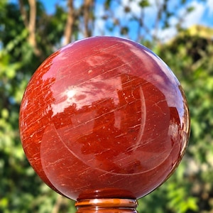 May include: A polished, deep red spherical stone with visible natural veining and a glossy, reflective surface. The sphere is mounted on a small, circular, tiered brown wooden stand, providing a stable base for the round mineral object.