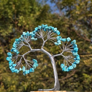 Może przedstawiać: Dekoracyjna rzeźba drzewa z drutu ze srebrnym metalowym pniem i gałęziami ozdobionymi licznymi małymi, bębnowanymi kamieniami w kolorze jasnego turkusu. Drzewo jest osadzone na płaskiej, okrągłej, jasnej kamiennej podstawie.