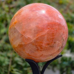 Op de afbeelding: Een gepolijste, bolvormige oranje en rode stenen bol met zichtbare interne kristallijne breuken en natuurlijke aders, rustend op een zwarte metalen driepootstandaard. Het oppervlak heeft een glanzende afwerking met warme tinten.