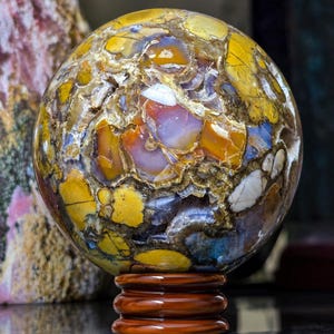 Op de afbeelding: Een gepolijste bolvormige steen met een complex brecciepatroon met levendige gele, doorschijnend oranje, dieppaarse en witte mineraalinklusionen. De bol staat op een gladde, donkerbruine, ronde houten displaystandaard.