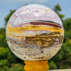 Op de afbeelding: Een gepolijste bolvormige steen met horizontale kleurstroken in dieprood, mosterdgeel, wit en grijs. Het oppervlak vertoont een complex, natuurlijk mineraalpatroon met gevarieerde texturen en insluitingen over de gehele bolvormige vorm.